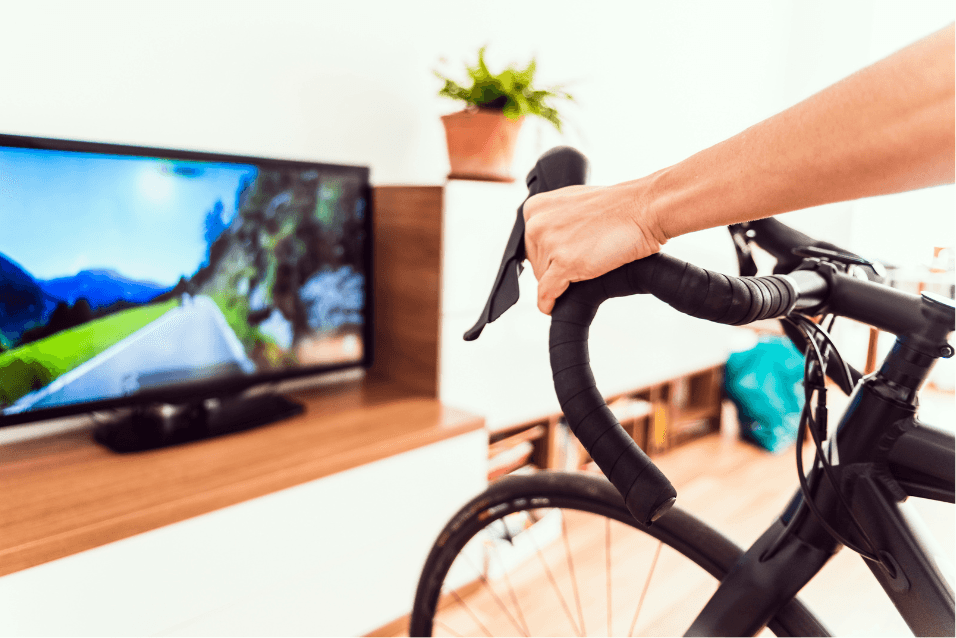 Cyclist's hand on road bike handlebars indoors, facing a TV showing a virtual road